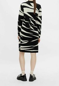 Robe en maille avec un motif en zigzag noir et blanc, manches longues, encolure ronde, et longueur au genou. Associée à des chaussures noires et des chaussettes crème.