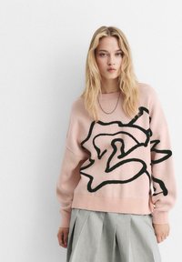 Maglione rosa chiaro con un design astratto nero, texture a maglia e polsini a coste. Abbinato a una gonna grigia plissettata.