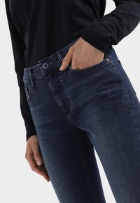 Dunkelblaue Jeans aus Denim mit einer figurbetonten Passform. Sie verfügen über ein klassisches Fünf-Taschen-Design, sichtbare Nähte und einen Knopfverschluss an der Taille.