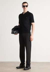Czarna krótka koszulka polo, dopasowany krój; czarne spodnie z wysokim stanem; trzyma czarną, teksturowaną torbę; czarne buty na platformie, elegancki design.