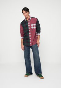 Kariertes Hemd mit roten und grünen Abschnitten sowie einer Brusttasche. Kombiniert mit locker sitzender, dunkler Jeans und grünen Sneakers.