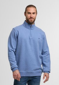 Šviesiai mėlynas zip-up džemperis su aukštu apykakle, ilgomis rankovėmis ir minkšta tekstūra. Priekyje yra mažas logotipas ant krūtinės. Dėvimas su džinsais.