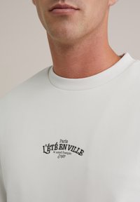 White cotton sweatshirt with a ribbed neckline. Features black embroidered text: "Paris L'ÉTÉ EN VILLE le soleil français 1987."