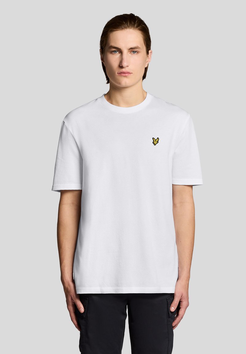 Νεαρός άνδρας με ανοιχτόχρωμο δέρμα και καστανά μαλλιά, φοράει λευκό κοντομάνικο πουκάμισο με μικρό κίτρινο λογότυπο πουλιού στο στήθος, στέκεται μπροστά σε γκρι φόντο.