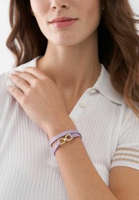 Purple leather bracelet with a gold clasp, featuring a double band design and a smooth texture. Worn on a wrist with a white polo shirt.