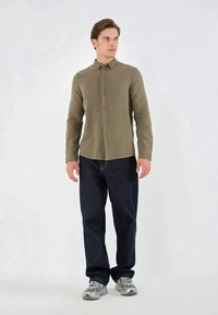 Uomo in piedi di profilo leggermente a destra, indossando una camicia a bottoni oliva, jeans scuri e sneakers grigie su sfondo bianco.