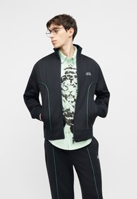 Fiatal férfi fekete, cipzáras dzsekiben és nadrágban, türkiz varrással, világoszöld gombos ing alatt fekete grafikával.