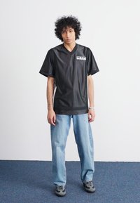 Camisa atlética preta com decote em V, com mangas curtas e um logótipo no peito, combinada com uns jeans largos azuis claros e sapatos cinzentos.