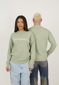 Lysegrøn strikket sweater med et "LEGACY" logo i creme, ribbede manchetter og kant. Modellen viser front- og bagview, parret med lyseblå jeans.