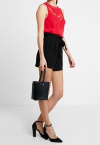 Haut sans manches rouge avec détail en dentelle, shorts noirs taille haute avec lien, mini sac structuré noir et chaussures à talons noirs.