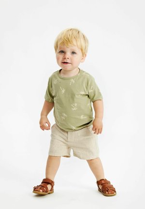 Camiseta verde claro con estampado de palmera, combinada con pantalones cortos beiges. Lleva sandalias marrones de tiras. El niño está de pie con las manos a los lados.