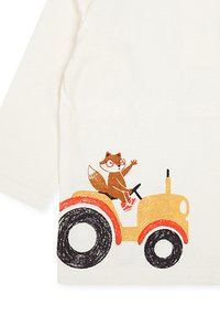 Un renard portant des lunettes et des chaussures rouges conduisant un tracteur jaune avec de grandes roues noires et rouges sur un t-shirt à manches longues blanc.