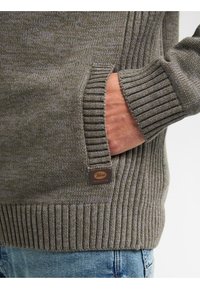 Grauer gestrickter Pullover mit geripptem Saum und Bündchen, ausgestattet mit einer Seitentasche und einem Leder-Logo-Patch. Strukturiertes Material verleiht Tiefe.