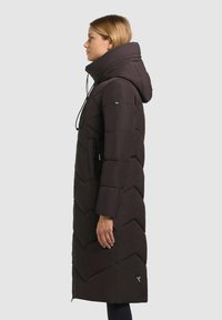 Lang puffercoat i mørkebrun med chevronmønster, høj krave og lynlåslommer på siden. Har justerbare snoretræk og logo-accent.