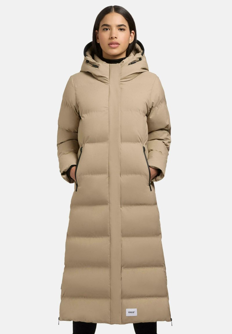 Lange beige pufferjas met een capuchon, voorzien van horizontale gestikte secties, ritszakken aan de zijkanten en een wit logo-label dichtbij de zoom.