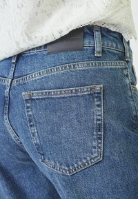 Blå denimjeans med en klassisk femficksdesign och en bakre läderlapp. Tyget har en urtvättad textur med synliga sömdetaljer.