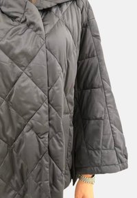 Manteau gris matelassé avec capuche, présentant un motif en losanges, des manches larges et un tissu doux et texturé.