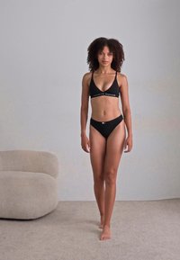 Čierny bikiny set s trojuholníkovým topom a vysokým pásom. Materiál vyzerá hladko, s bielym logom pozdĺž horného pásu.