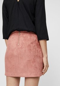 Mini-jupe en daim rose poussiéreux avec une texture lisse et une fermeture éclair métallique à l'arrière. Associée à un haut noir, mettant en valeur un design simple.
