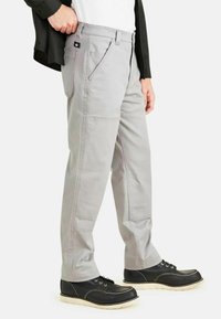 Pantalons cargo gris clair avec une coupe droite, présentant des coutures détaillées et un écusson noir à l'arrière de la poche. Portés avec des chaussures noires à lacets.