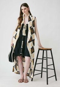 Long black dress with a sleeveless, patterned cream overlay featuring floral designs. Model wears brown slide sandals and holds a bag.