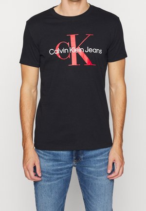 Homme portant un t-shirt noir avec un grand logo Calvin Klein Jeans rouge et blanc et un jean bleu sur un fond uni.