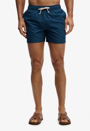 Mężczyzna w niebieskich kąpielówkach z białym sznurkiem i brązowych skórzanych sandałach, stojący na białym tle.