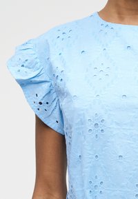 Φωτεινό γαλάζιο φόρεμα με κεντητό λουλουδάτο σχέδιο με ανοίγματα και φαρδιά μανίκια σε πρόσωπο με μέτριο τόνο δέρματος.