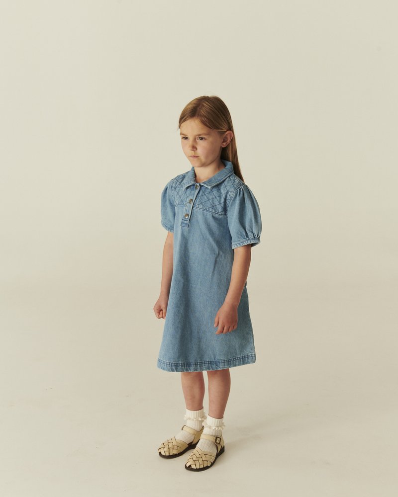 Junges Mädchen in einem hellblauen Jeanskleid mit Puffärmeln und beigen Sandalen mit weißen Socken, stehend vor einem einfarbigen beigen Hintergrund.