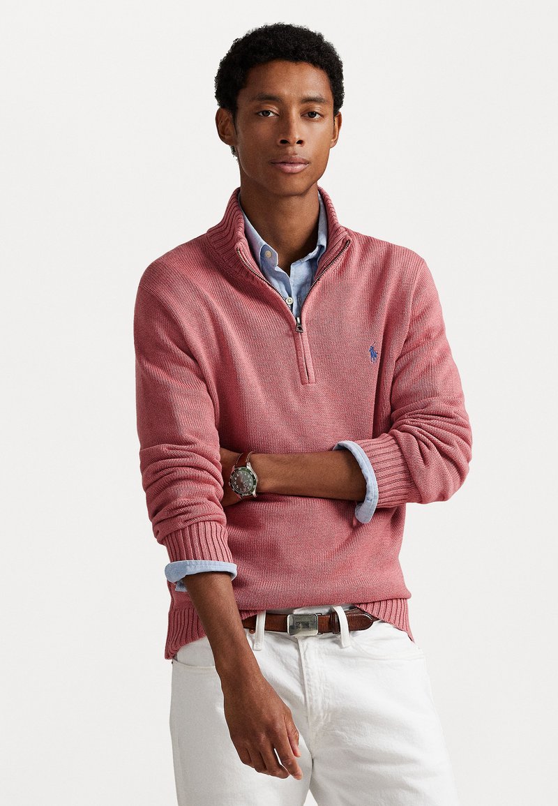 Κόκκινο πουλόβερ με φερμουάρ και ραμμένες μανσέτες και στρίφωμα, σε επικάλυψη με ανοιχτό μπλε πουκάμισο με κολάρο, συνδυασμένο με λευκά παντελόνια και ασημένιο ρολόι.