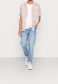 Koszula z krótkim rękawem w jasnobeżowy wzór, noszona na białej koszulce, w połączeniu z jasnoniebieskimi dżinsami i białymi tenisówkami, stojąca na jednolitym tle.