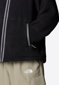 Svart fleecejacka med kontrasterande silverblixtlås och fickdetaljer, med texturerad tyg. Matchas med beige byxor som visar en logotyp.