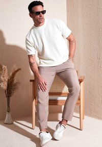 Hvid ribbet kortærmet skjorte, beige slim-fit bukser, hvide og grønne sneakers, siddende på en træstol med rattan detaljer, lys baggrund.