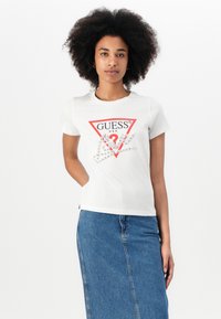Maglietta in cotone bianco con un logo triangolare rosso che presenta la scritta "GUESS?" e grafica delle mani con strass. Vestibilità casual, maniche corte, colletto rotondo.