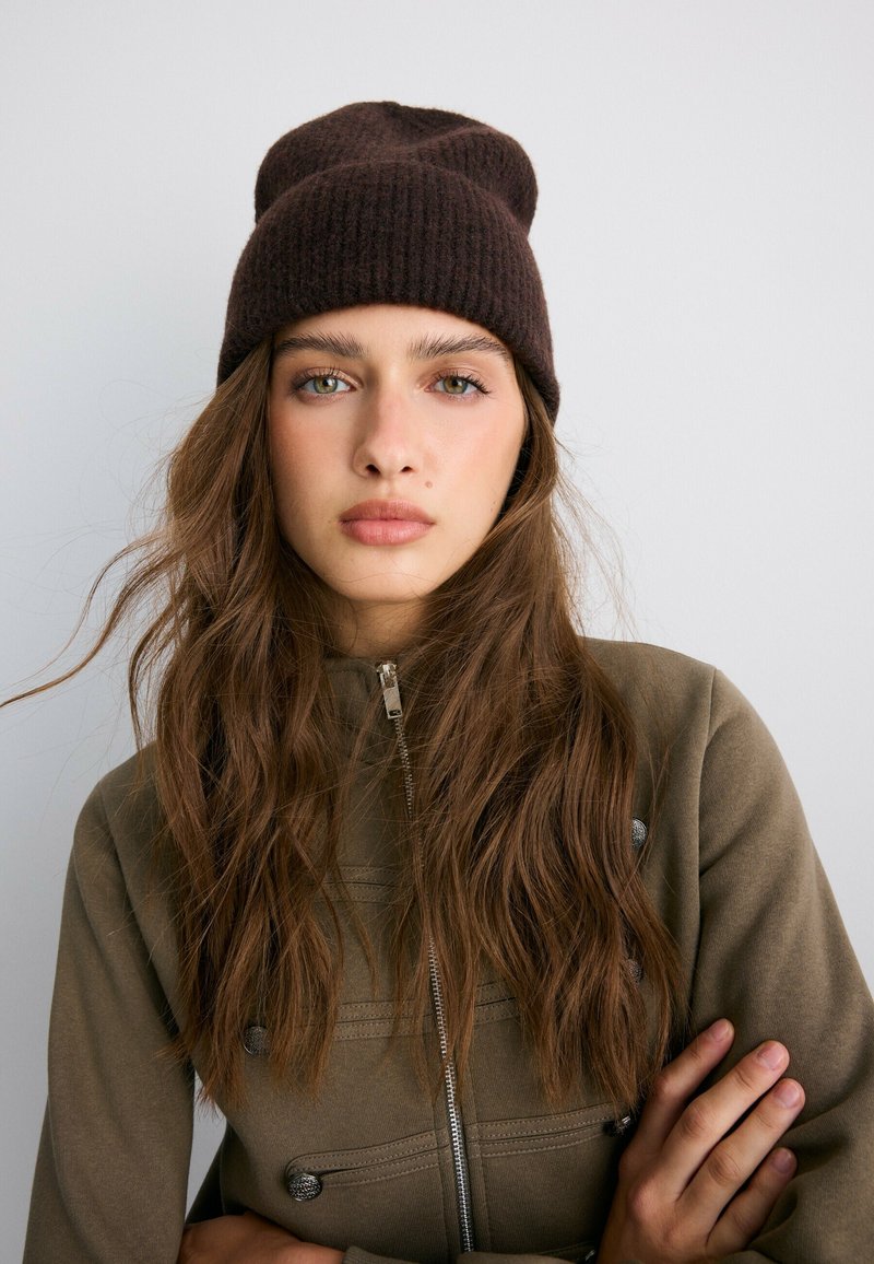 Jeune femme aux longs cheveux bruns portant un bonnet en tricot marron foncé et une veste vert olive, regardant directement avec une expression neutre.
