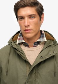 Olive green jacket with a hood, featuring a fuzzy collar. Worn over a beige sweater and a plaid shirt with red and navy tones.