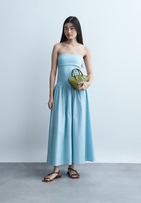 Νεαρή γυναίκα σε αμάνικο ανοιχτό μπλε φόρεμα, κρατώντας μια μικρή τσάντα ελιάς, φορώντας καφέ σανδάλια, στέκεται μπροστά σε ανοιχτού χρώματος τοίχο.