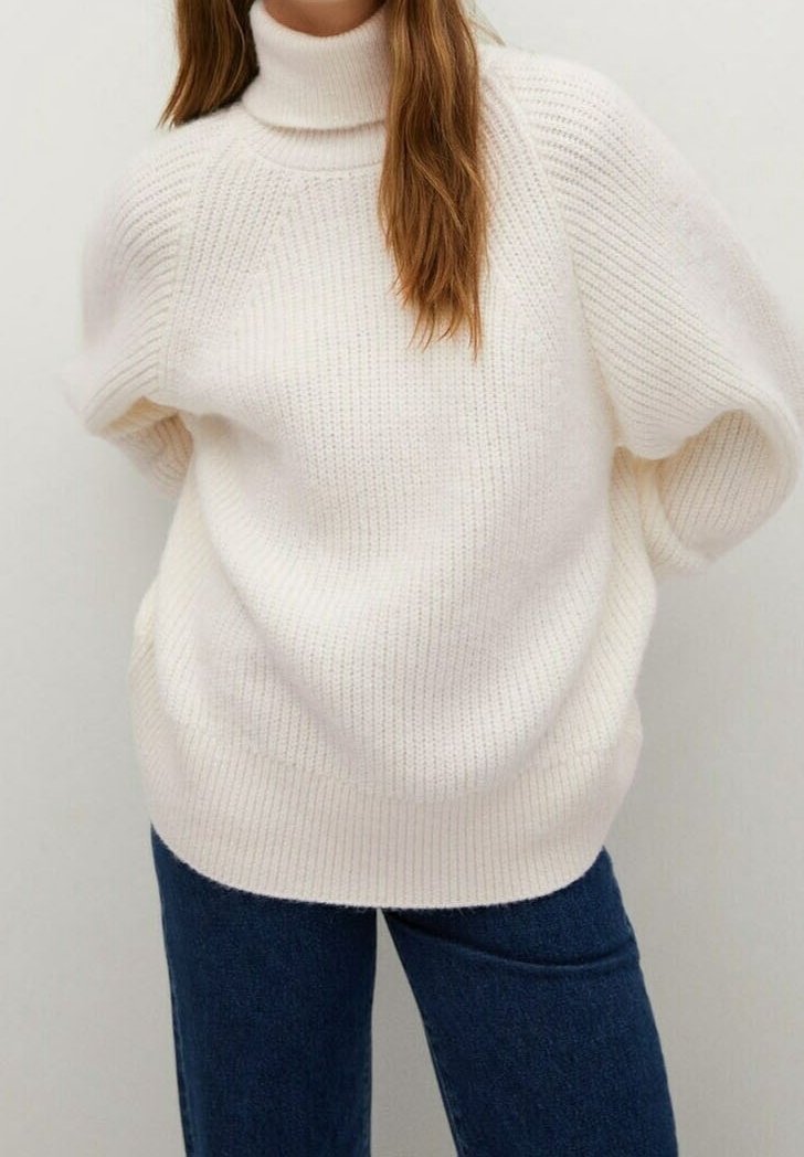 Gruby golf w miękkim, białym dzianinowym materiale, o luźnym kroju i prążkowanej fakturze, zestawiony z ciemnoniebieskimi dżinsami.