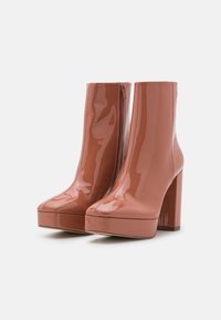 Botas de tobillo de charol marrón brillante con plataformas y tacones altos gruesos, con cremalleras laterales, sobre un fondo blanco.