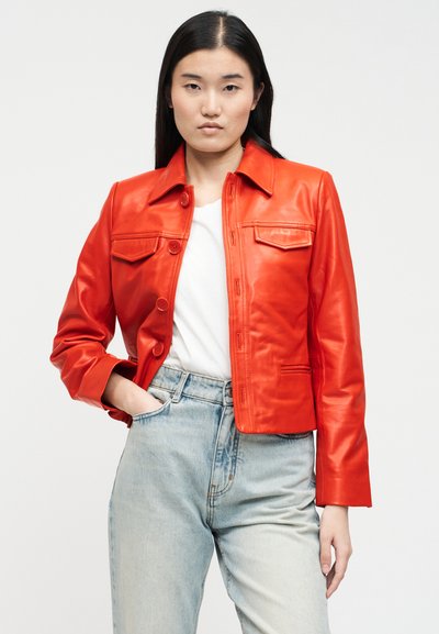 Γυναίκα με φωτεινό κόκκινο δερμάτινο μπουφάν με κουμπιά πάνω από λευκό πουκάμισο και ανοιχτό μπλε ψηλόμεσο τζιν, με το χέρι στην τσέπη, ουδέτερο φόντο.