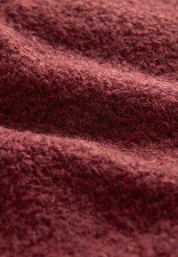 Gros plan d'un tissu en laine doux, texturé, de couleur bordeaux profond avec des fibres bouclées et des plis subtils créant une apparence conviviale et chaleureuse.