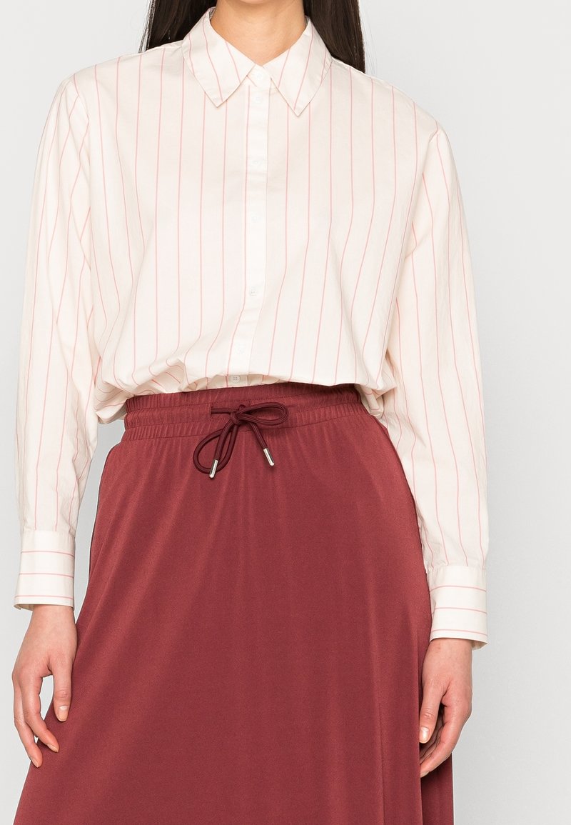 Femme portant une chemise blanche à manches longues avec de fines rayures verticales rouges, rentrée dans une jupe bordeaux taille haute avec un cordon à la taille.