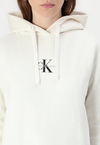 Balta pūkinė striukė su priekinę kišenę ir šnurine kapišonu. Yra juodas "CK" logotipas ir žemiau jo tekstas "Calvin Klein Jeans". Minkšta audinio tekstūra.