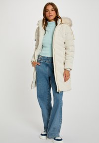 Polstret vinterjakke i lys beige med hætte med pelskant, med en for-lynlås og sidelommer, båret over en mintgrøn top og blå jeans.