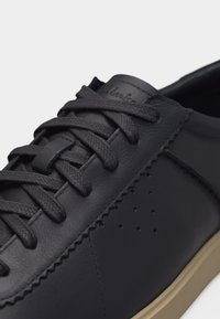 Μαύρο δερμάτινο αθλητικό παπούτσι με μαύρα κορδόνια, πλευρικές οπές αερισμού με διάτρηση και μπεζ σόλα, σε κοντινή λήψη με λεπτομέρεια.