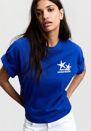 Mladá žena s dlhými tmavými vlasmi, oblečená v modrom tričku s vyhrnutými rukávmi a bielym textom a grafikou hviezdneho kraba na hrudi, pózuje s rukami v bok.