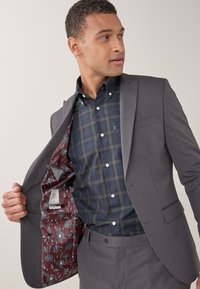 Giacca grigia con una fodera floreale borgogna, indossata sopra una camicia a scacchi scuri. Presenta bottoni sulla camicia e un taglio su misura.
