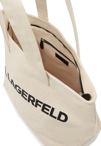 Sac fourre-tout en toile couleur crème avec texte noir. Comprend une poche intérieure zippée et des bretelles solides, mettant en avant un design minimaliste.