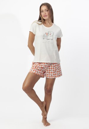 Mujer con camisa blanca de manga corta con un oso leyendo en un sillón y pantalones cortos de cuadros naranjas con estampado de oso, de pie descalza.