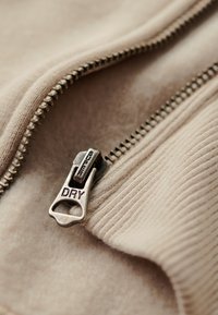 Prenda beige con cremallera y tejido texturizado. Presenta un tirador de cremallera metálico marcado como "SUPER DRY" y un acento acanalado a lo largo del borde.